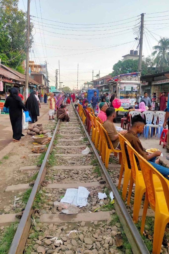 People sat on orange plastic chairs backed up to railway line, markets stalls either side.