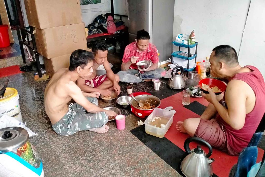 Four men sit barefoot on polished floor eating food from bowls.