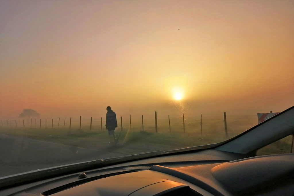 View from car dashboard of walking man silhouetted on misty road at sunset.