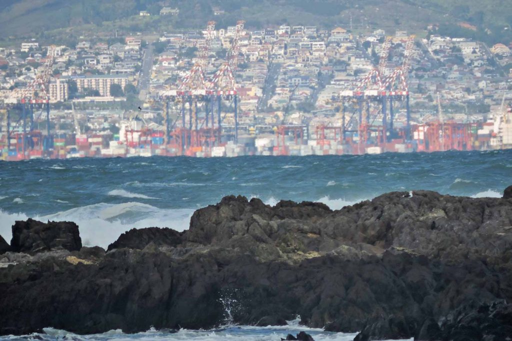 Waves breaking on rocks in foreground with colourful containers and cranes on the horizon.