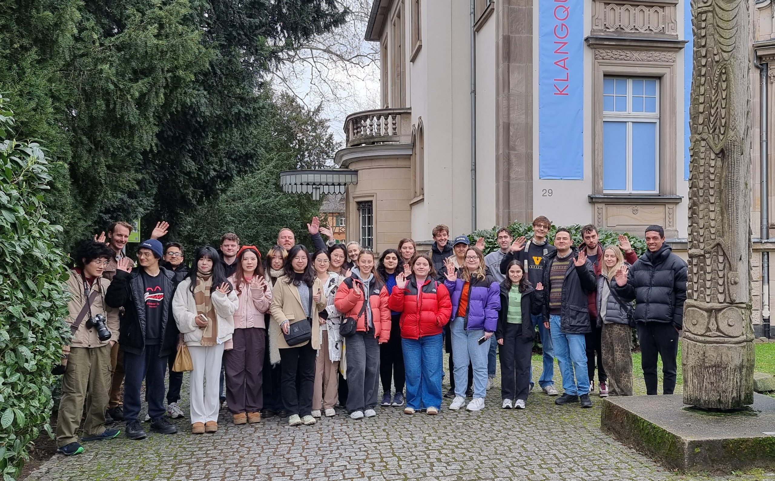 Group of students standing in a traditional garden surrounded by sculptural columns. An old house is behind them.